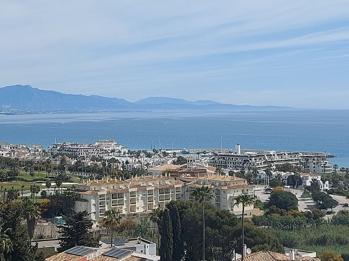 View of a coastal town with apartment buildings, a marina, and mountains in the background overlooking the sea.