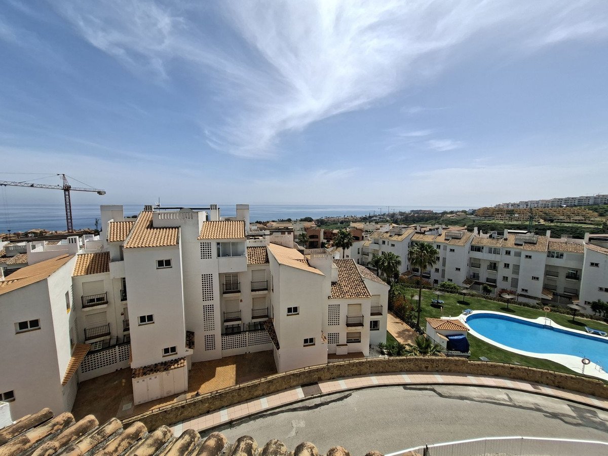 View of a residential complex with white buildings, balconies, a swimming pool, and the sea in the distance under a blue sky.