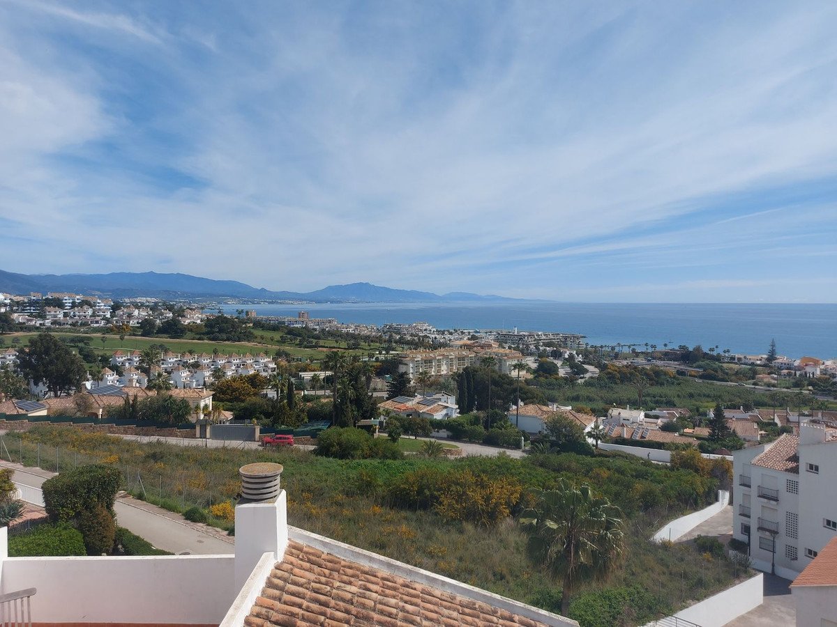 Expansive sea and town view with mountains in the distance under a cloudy blue sky.