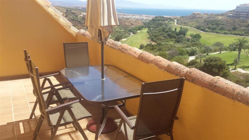 A terrace with a glass table, chairs, and umbrella, overlooking a golf course and the sea.