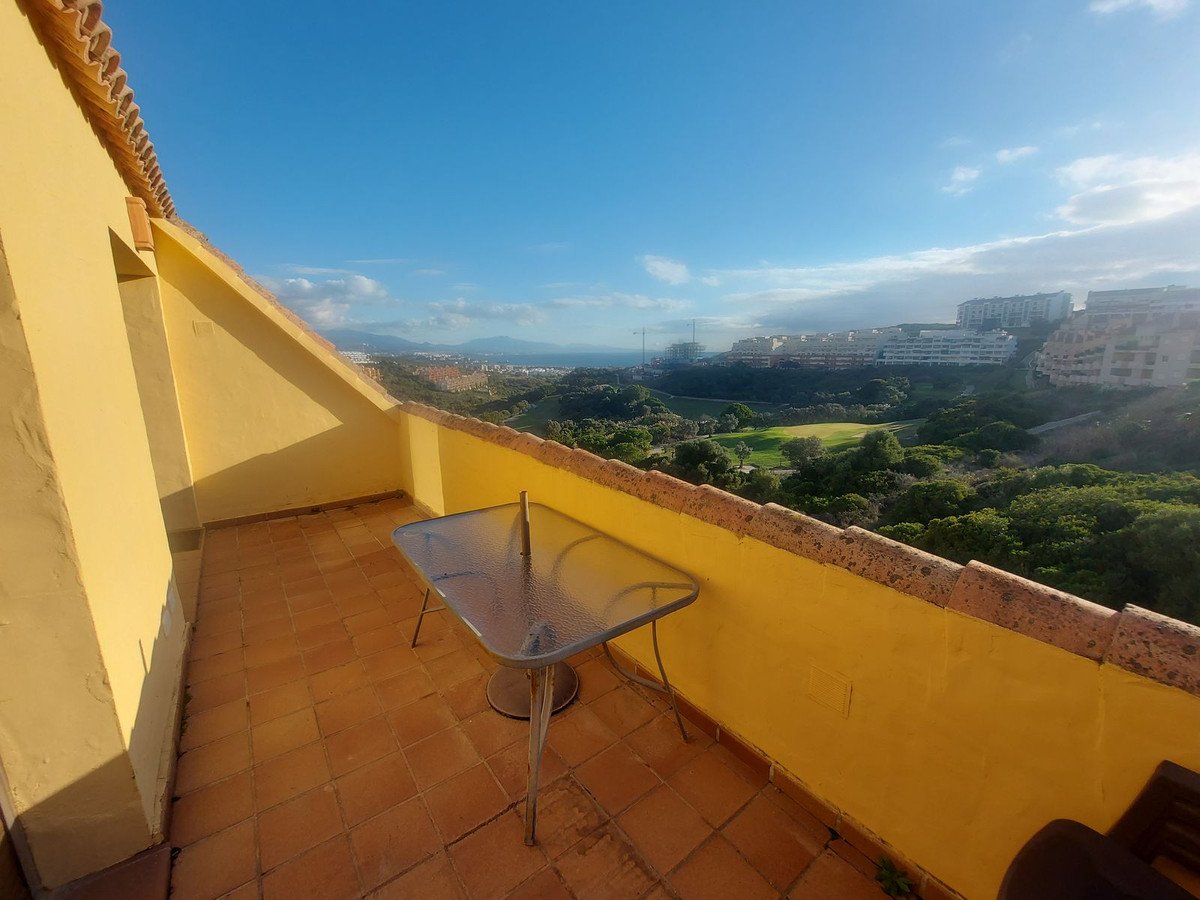 Terrace with glass table overlooking a golf course, distant sea, and buildings under a blue sky.