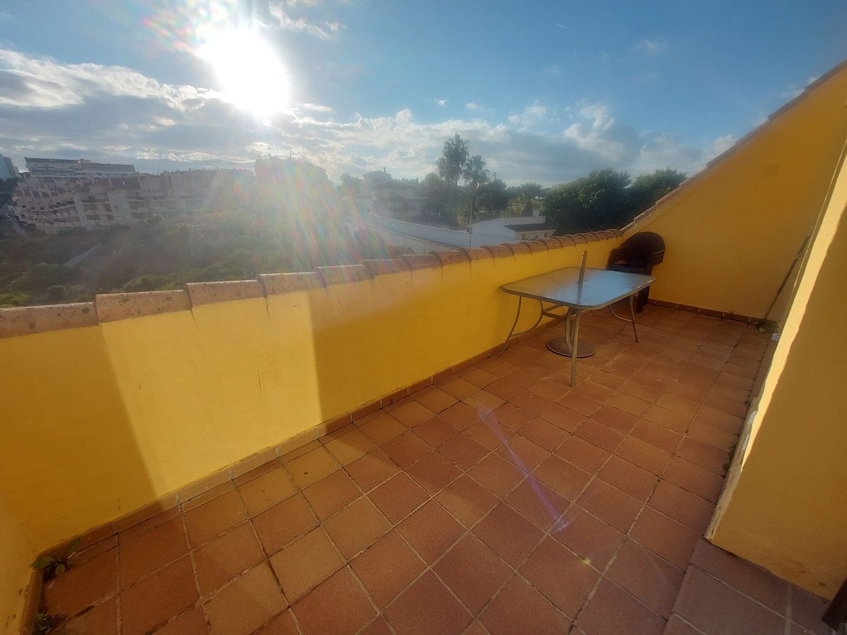 Sunny terrace with a table and chair, overlooking buildings and greenery under a bright blue sky.