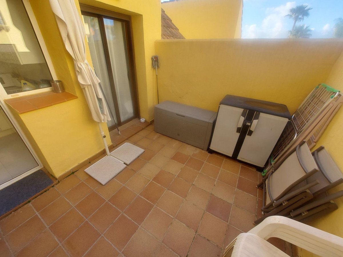 Terrace with storage boxes, folded chairs, and a closed umbrella against yellow walls under a blue sky with palm trees.