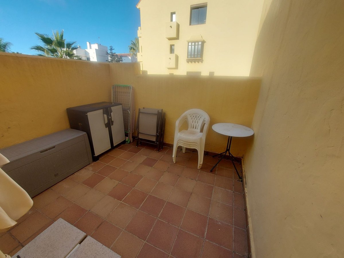 A small terrace with storage boxes, a chair, and a table, featuring palm trees and a building in the background.