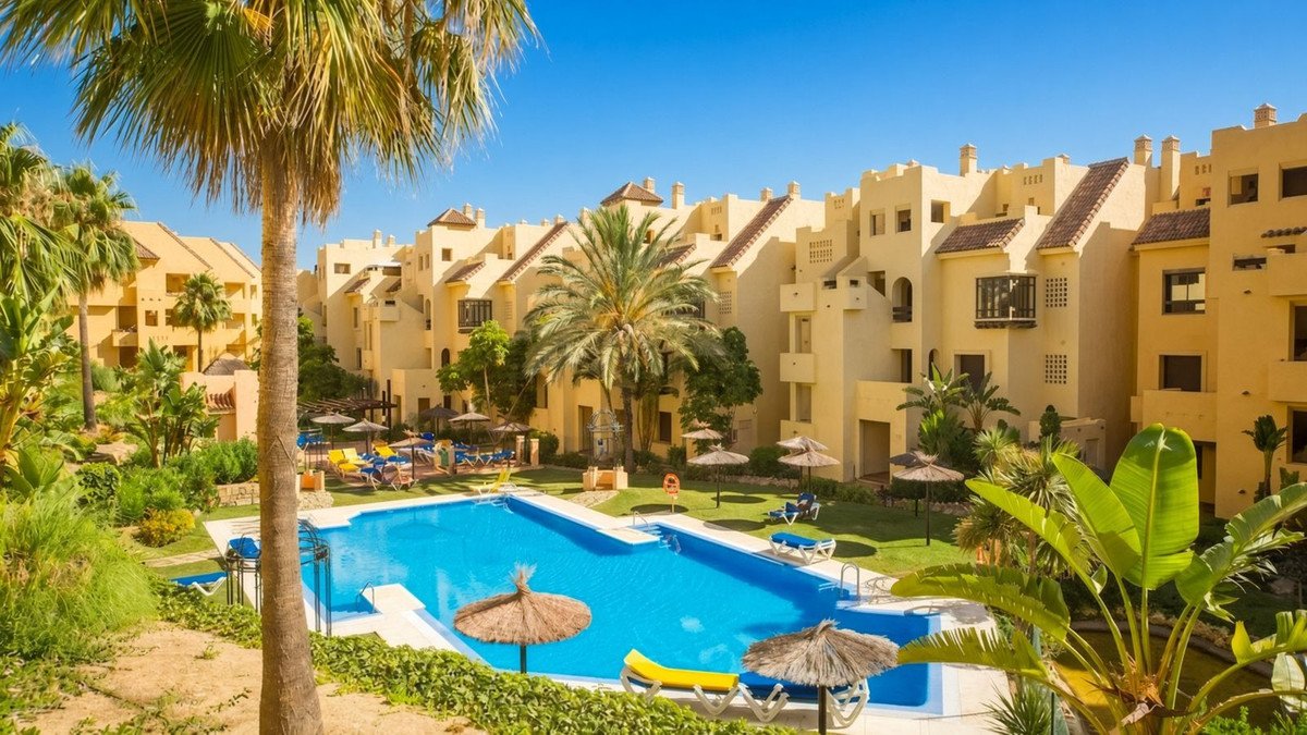 A sunny resort complex with a bright blue swimming pool, sun loungers, and palm trees against a clear sky.