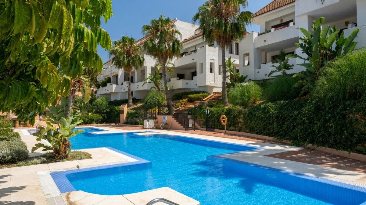 A large, bright blue swimming pool in front of a white apartment complex with palm trees and lush greenery under a clear blue sky.
