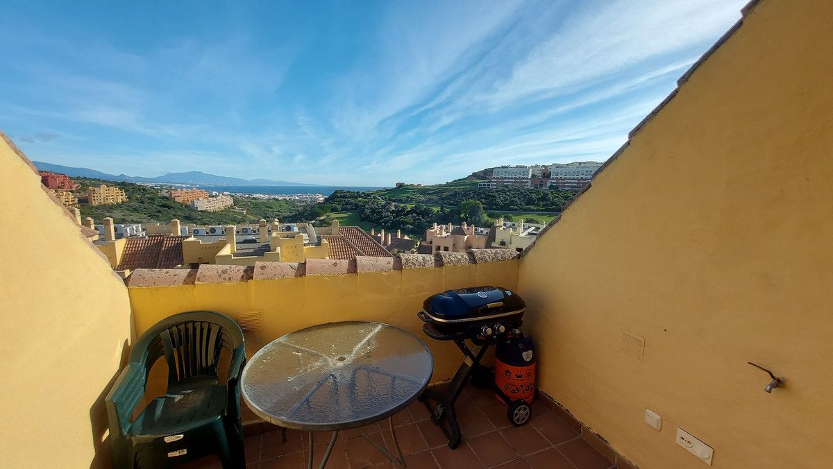 Terrace with a table, chair, and grill, overlooking a town and the sea under a blue sky.