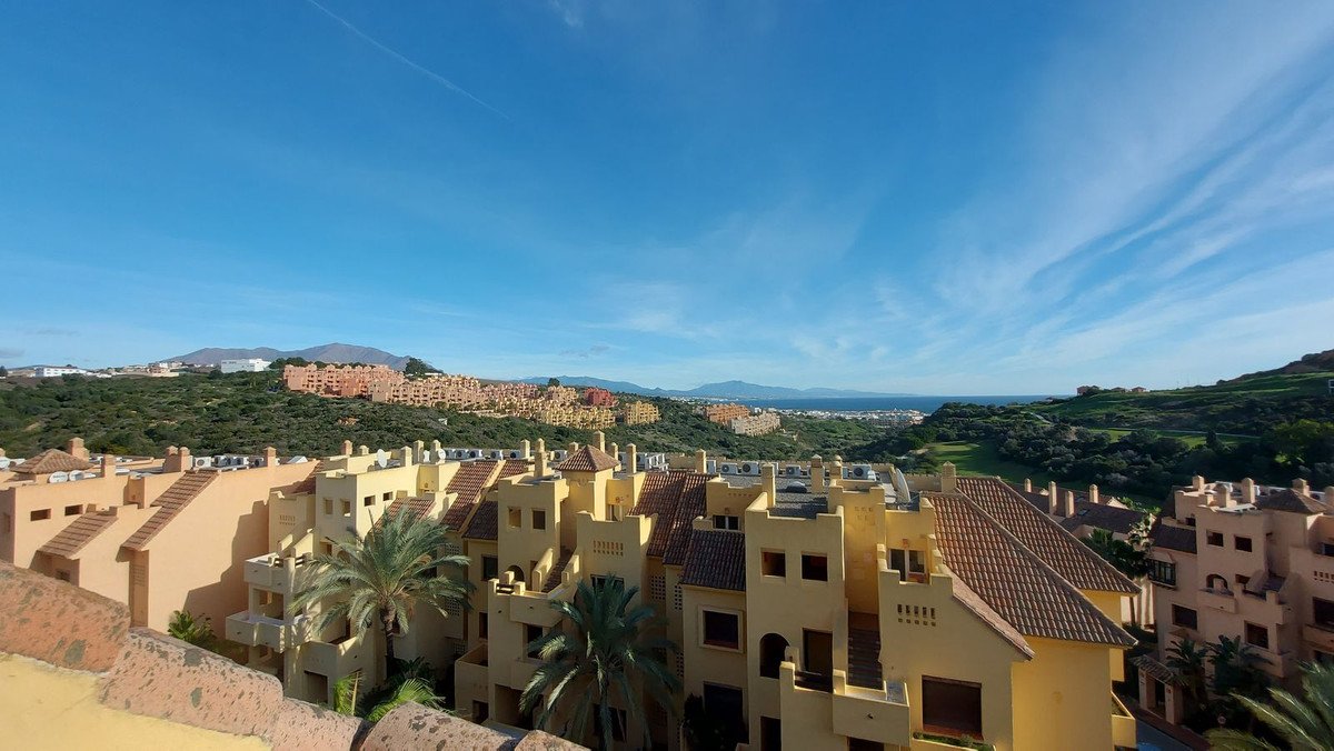 View from apartment balcony showing a complex of yellow buildings, green hills, and the sea in the distance under a blue sky.