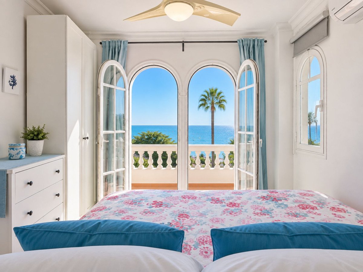 Bedroom with double doors opening to a balcony with sea view, palm tree, and railing.
