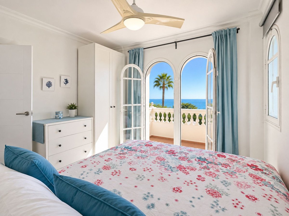 Bedroom with a floral comforter, blue curtains, and a balcony offering a sea view with a palm tree.
