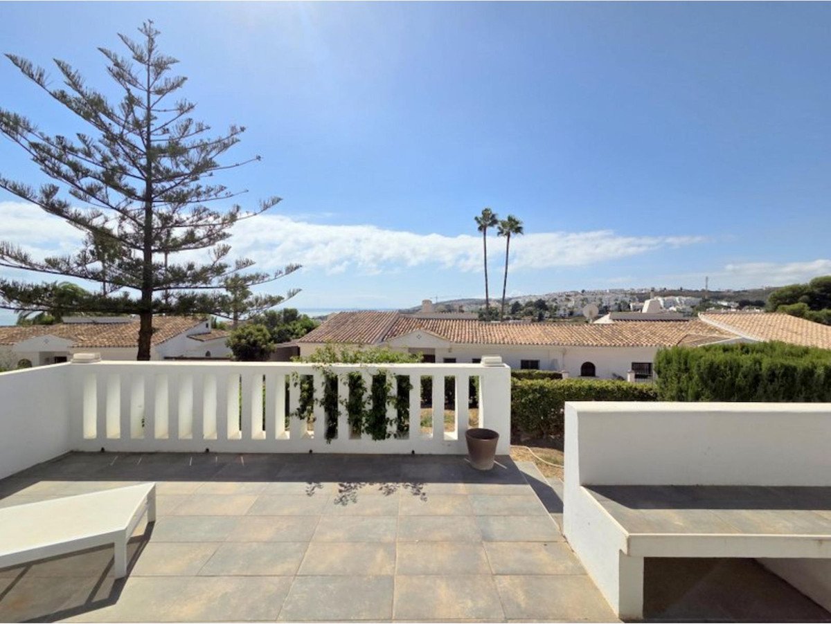 Terrace with white railing, built-in bench, overlooking rooftops, palm trees, and distant sea under a blue sky.