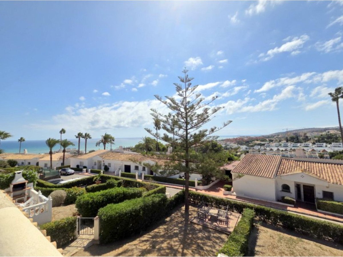 View from a terrace overlooking a garden, white houses, palm trees, and the blue sea under a sunny sky.