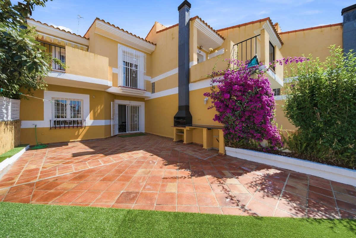 A sunny yellow house exterior with a tiled terrace, barbecue, and blooming purple bougainvillea in the garden.