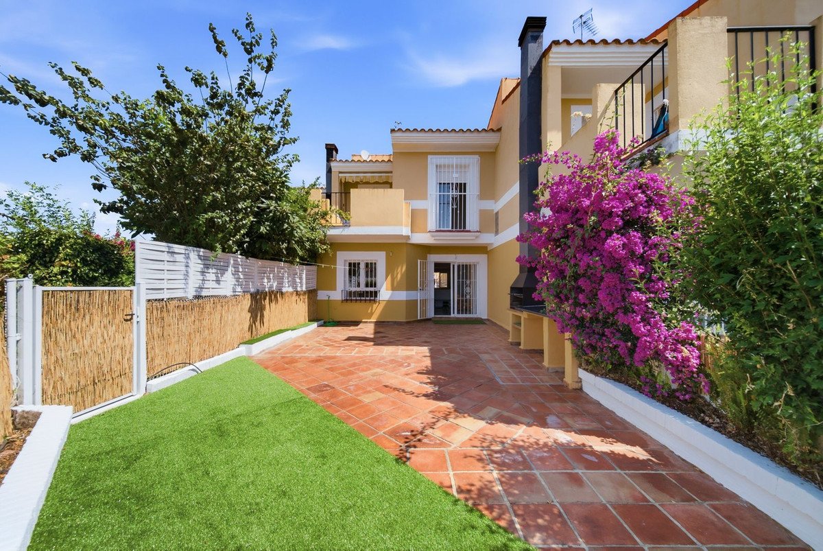 Yellow house exterior with a tiled patio, artificial grass lawn, BBQ grill, and purple bougainvillea flowers.