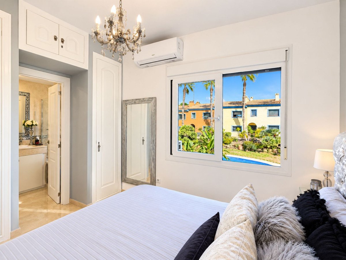 Bedroom with a view of palm trees, colorful houses, and a swimming pool through the window.