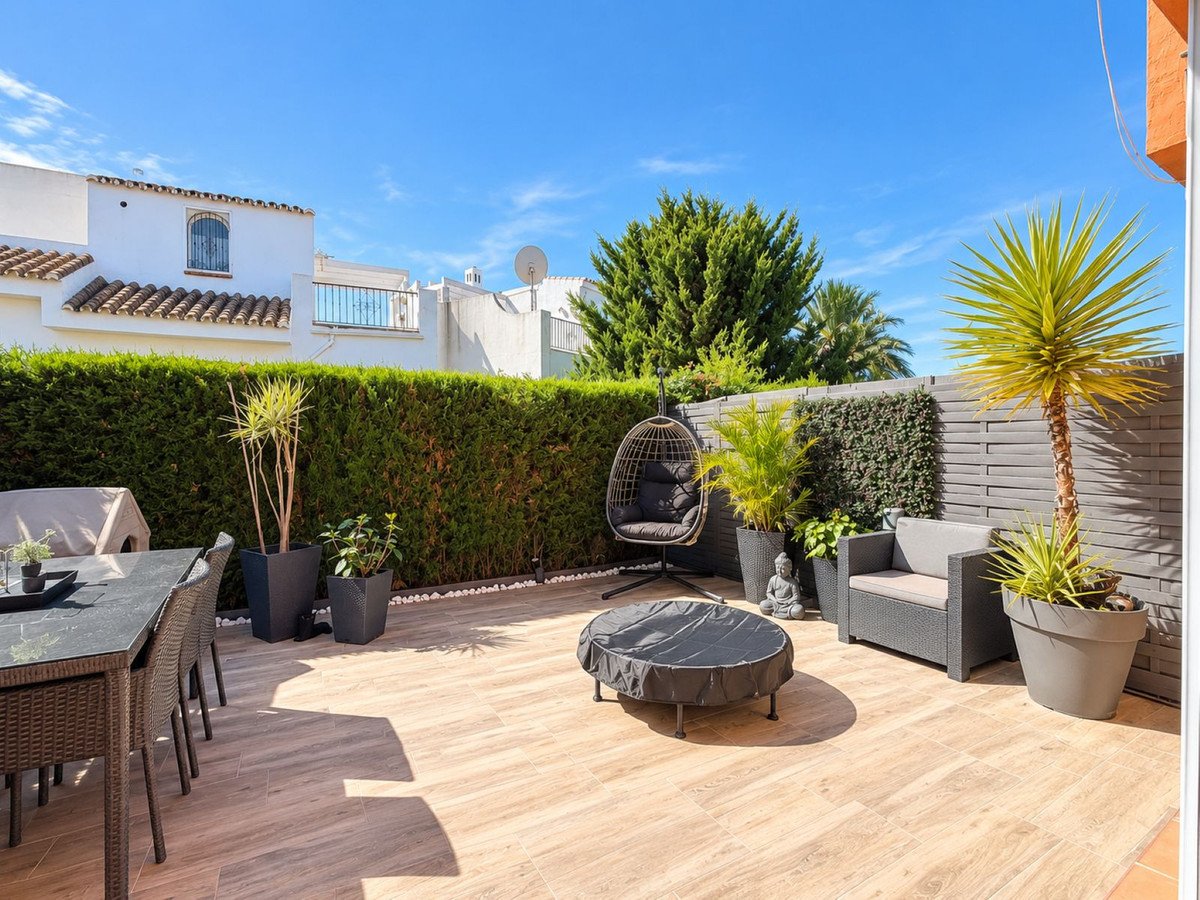 A paved terrace with outdoor seating, hanging chair, dining table, and lush green plants under a clear blue sky.
