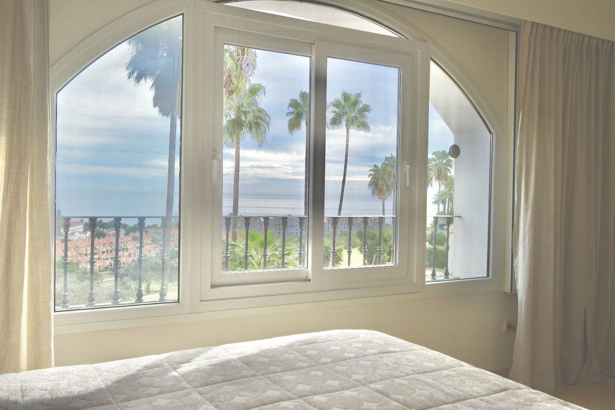 Bedroom with arched window and sliding doors offering a sea view, balcony with palm trees, and town below.