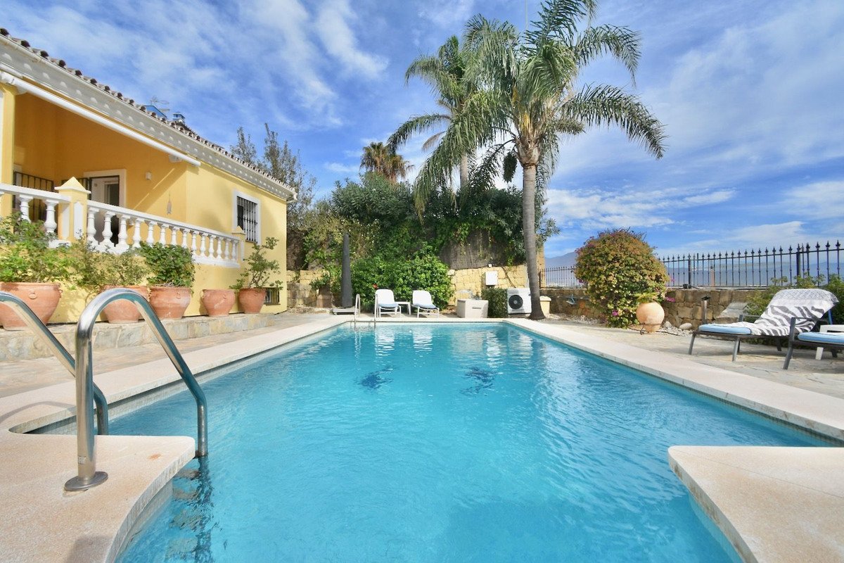 A bright yellow villa with a swimming pool and terrace, featuring lounge chairs, palm trees, and lush greenery under a blue sky.
