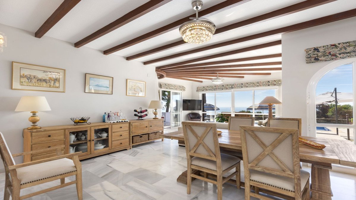Spacious living and dining area with wooden beams, sea view windows, and access to an outdoor terrace with a pool.