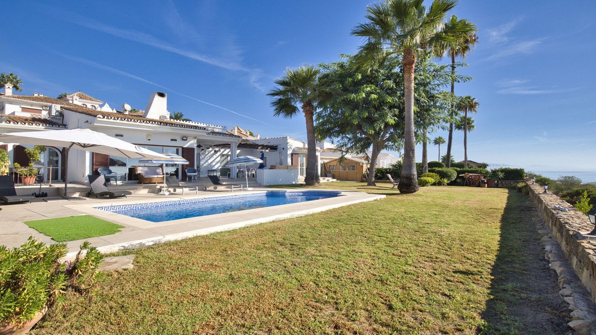 Spacious villa with a blue tiled swimming pool, lush green lawn, palm trees, and a distant view of the sea.