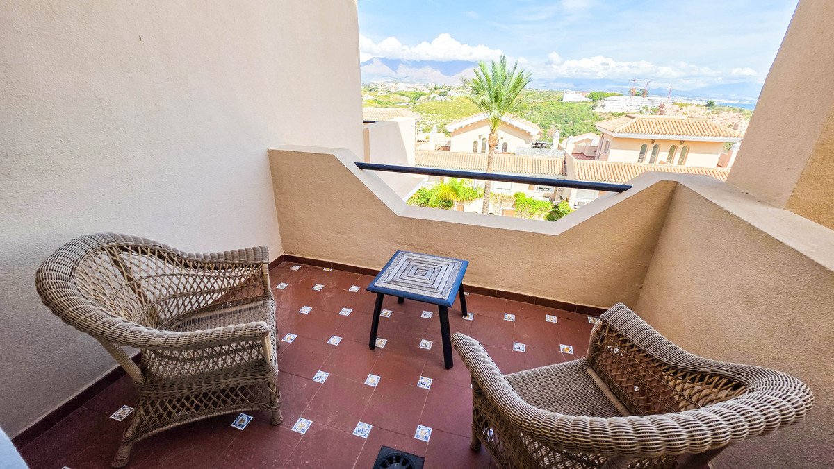 A balcony with two wicker chairs and a table, offering a distant view of the sea and mountains under a blue sky.