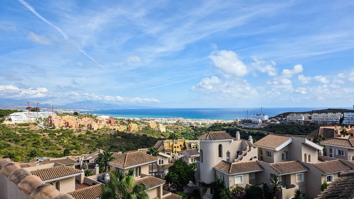 Panoramic view of a townhouse complex with terracotta roofs overlooking the sea and a distant town under a blue sky with clouds.