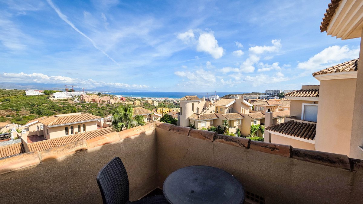 A balcony with a small table and chair offering a scenic view of the sea, rooftops, and distant hills under a blue sky.