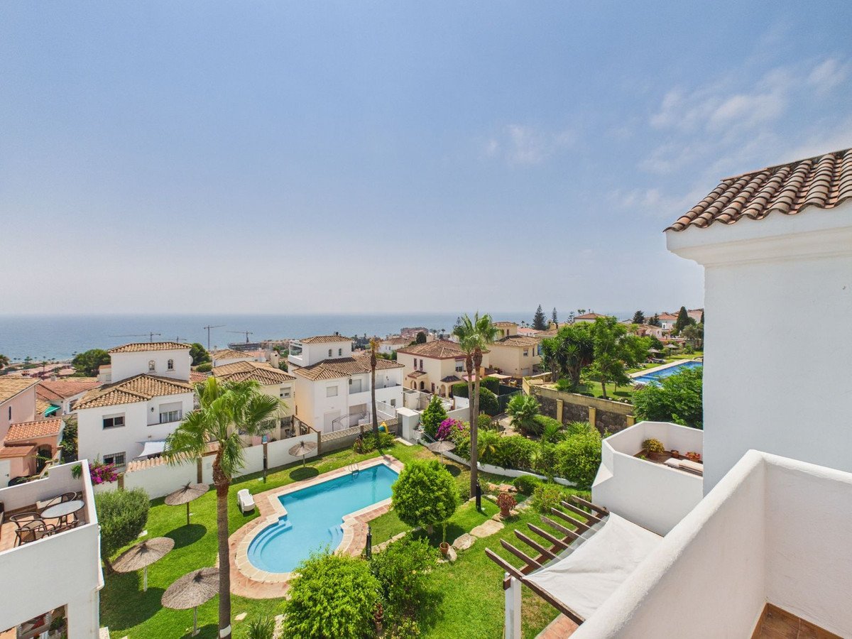 View from a balcony overlooking a swimming pool, gardens, houses, and the sea in the distance under a clear blue sky.