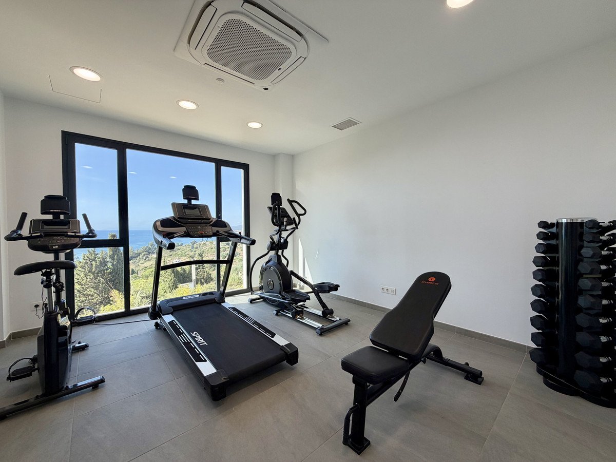 Home gym with treadmill, stationary bike, elliptical, weight bench, and dumbbells, featuring a large window with a sea view.