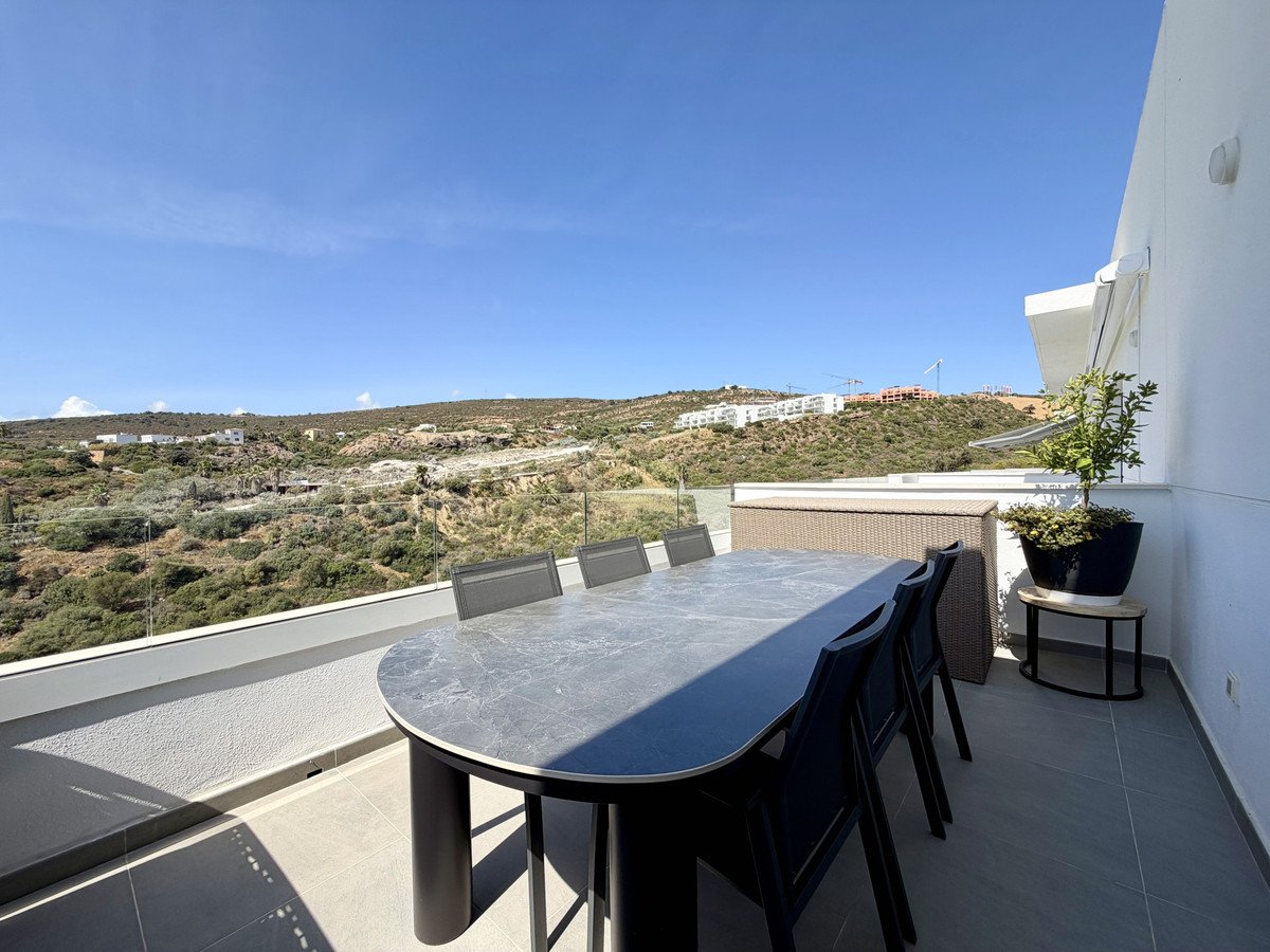 A terrace with a dining table and chairs, overlooking a landscape with distant buildings and hills under a clear blue sky.