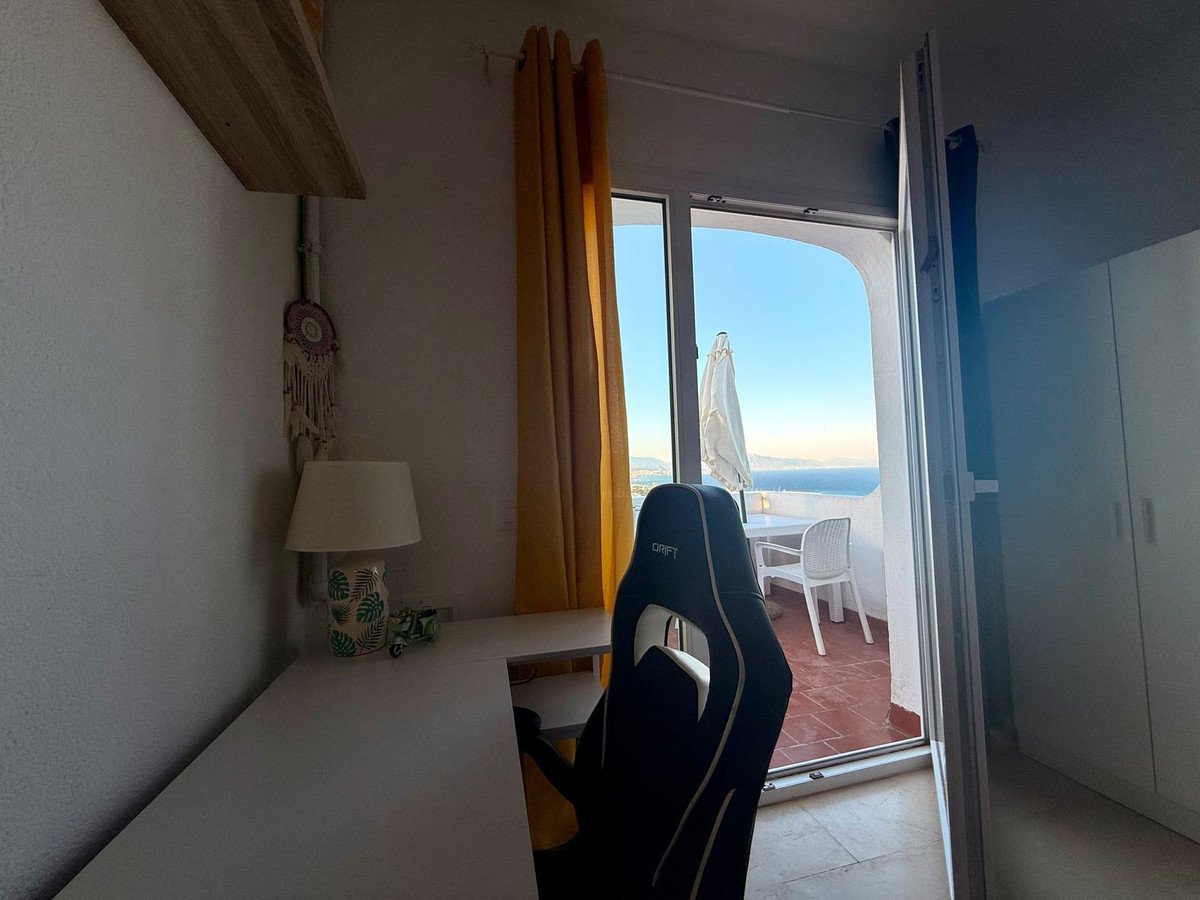 Bedroom with a desk and gaming chair, overlooking a balcony with a table, chairs, and a beautiful sea view.