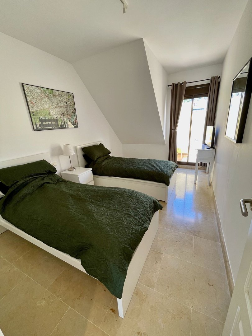 A bright bedroom with two green-covered beds, a sloped ceiling, and a glass door leading to a balcony.