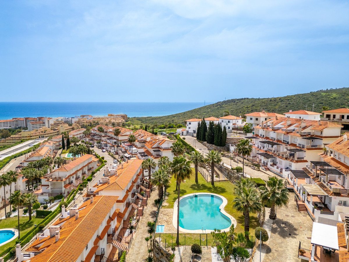 Aerial view of a residential complex with a pool, palm trees, and the sea in the background under a clear blue sky.