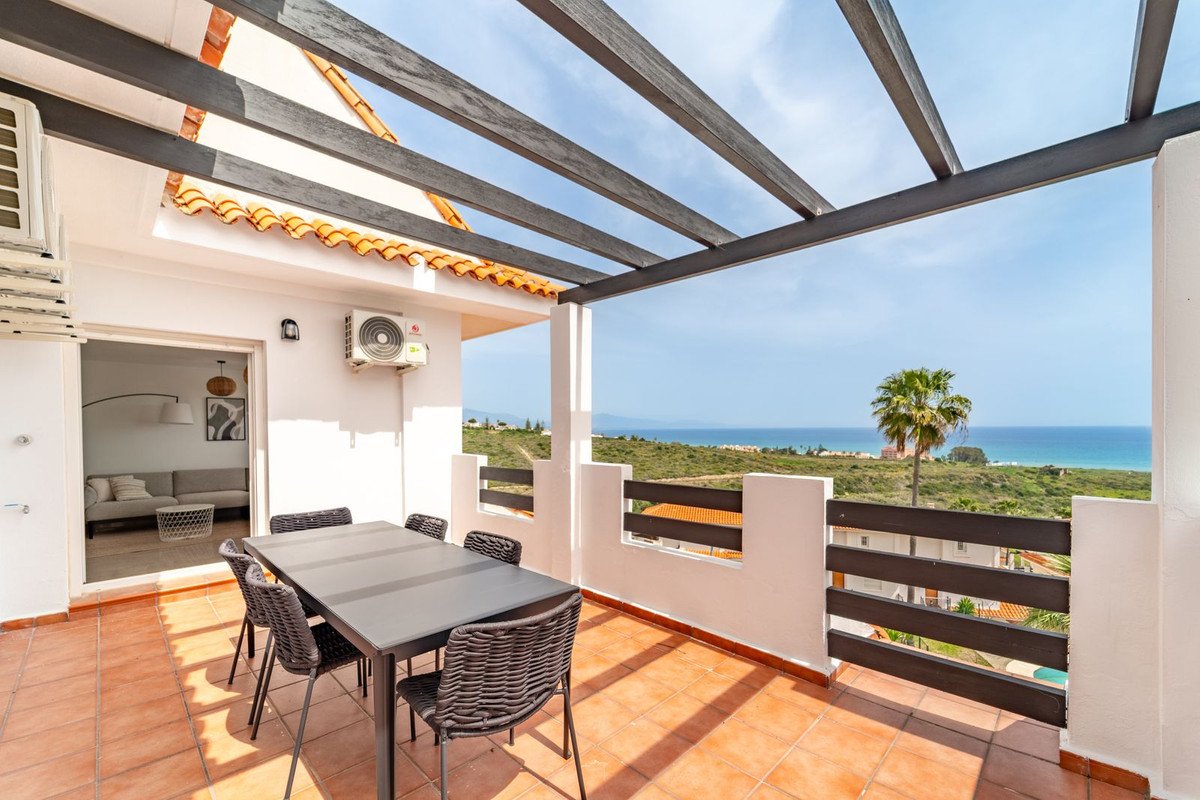 A covered terrace with a dining table and chairs offers a stunning sea view with a palm tree in the foreground.