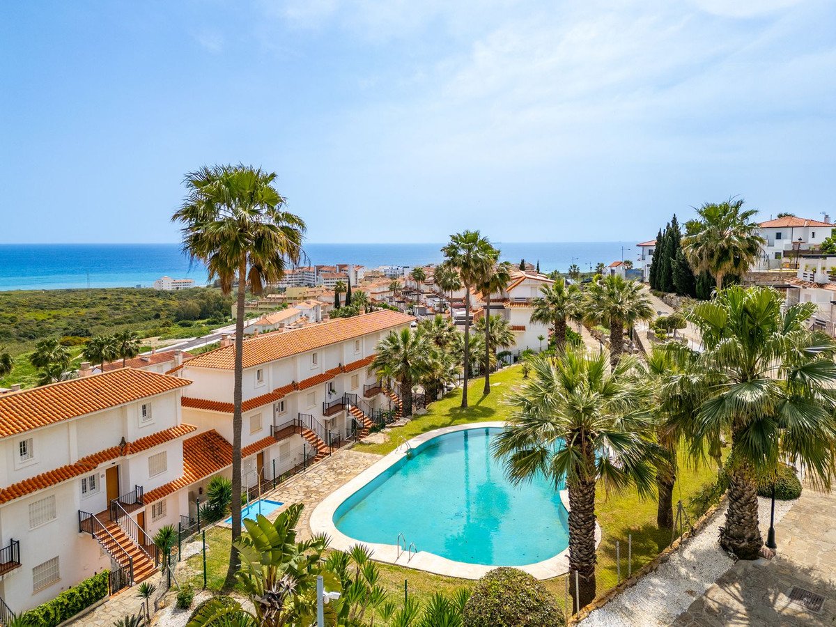 A resort complex with white buildings, a blue swimming pool, palm trees, and a distant sea view under a clear blue sky.