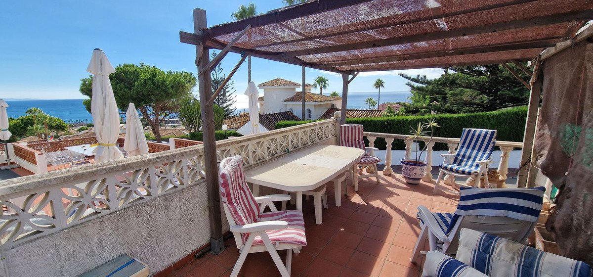 Covered terrace with dining table and chairs, overlooking the sea and surrounding rooftops under a clear blue sky.