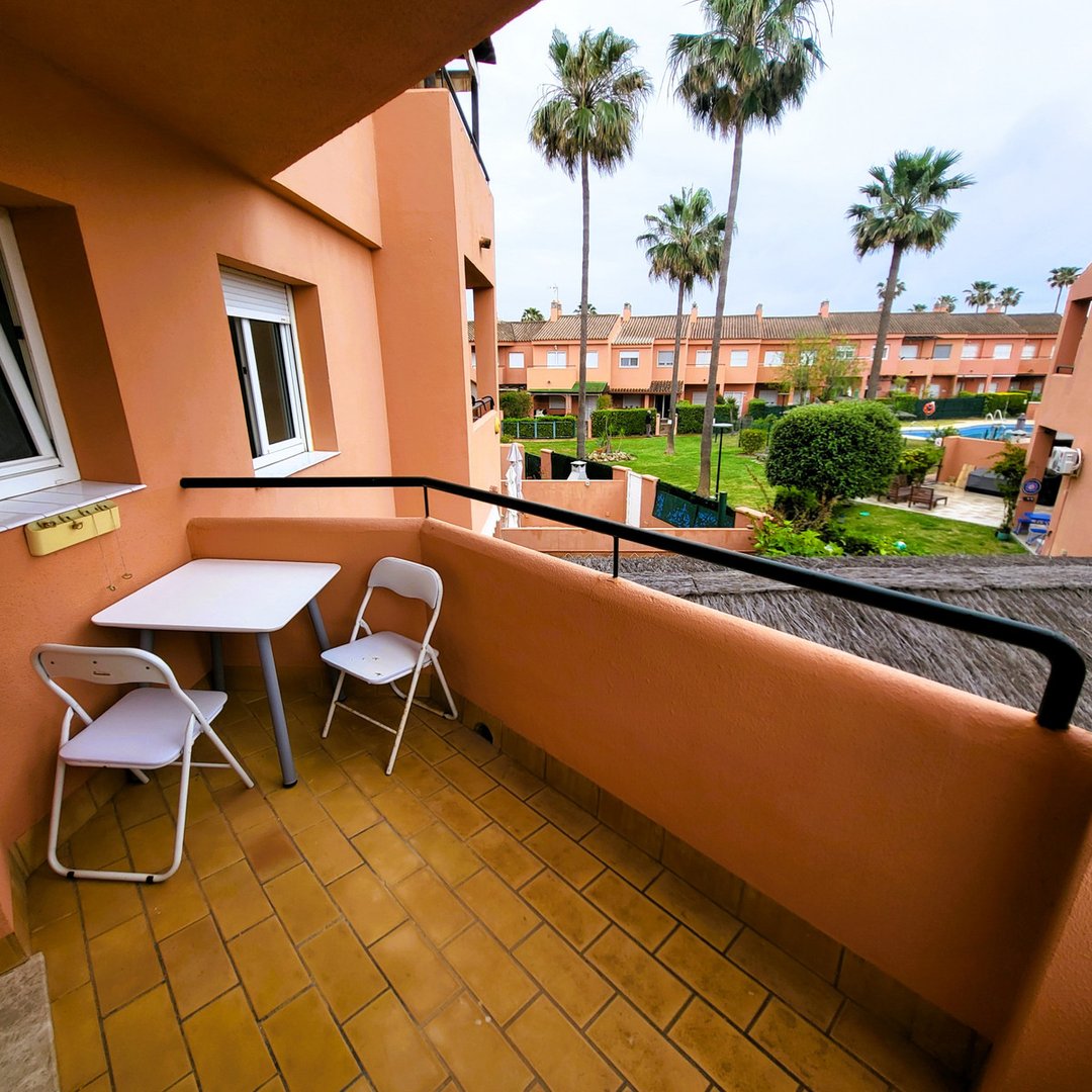 Balcony with table and chairs, orange building, palm trees, pool view.