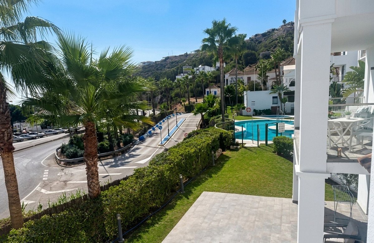 View from a balcony overlooking a swimming pool, garden, and palm trees with a hillside in the background.