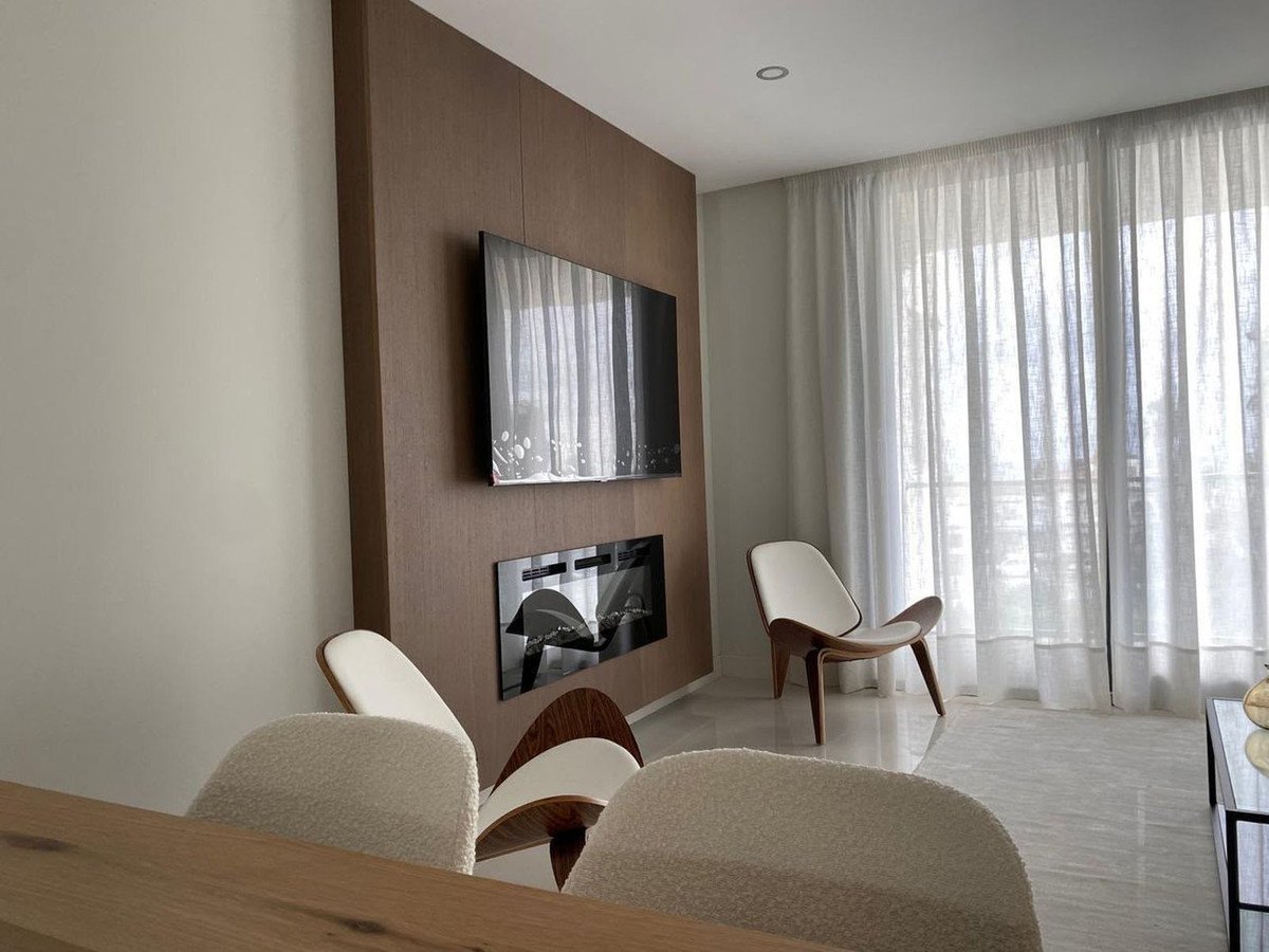Modern living room with a mounted TV, electric fireplace, and stylish chairs, with sheer curtains letting in natural light.