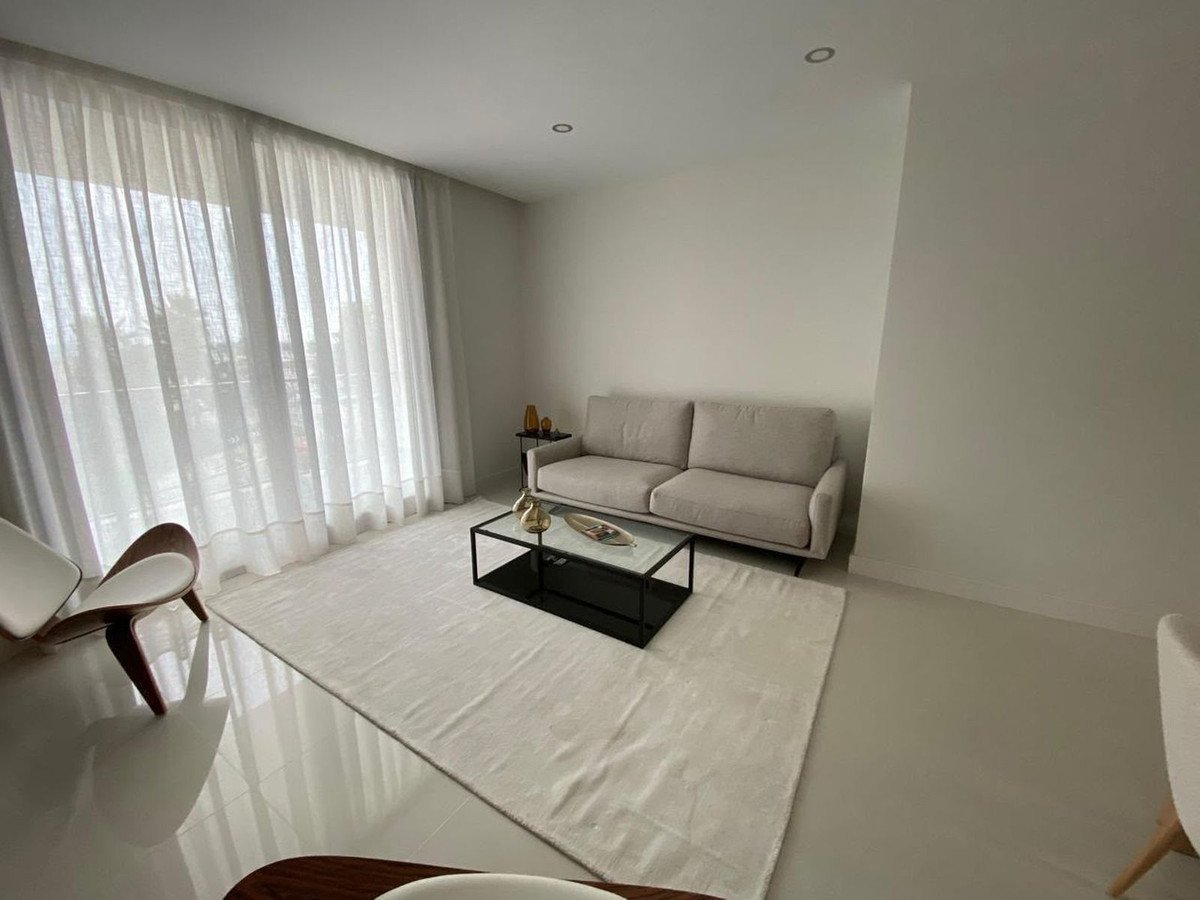 A modern living room with a beige sofa, black coffee table, white rug, and large sheer curtains revealing outdoor greenery.