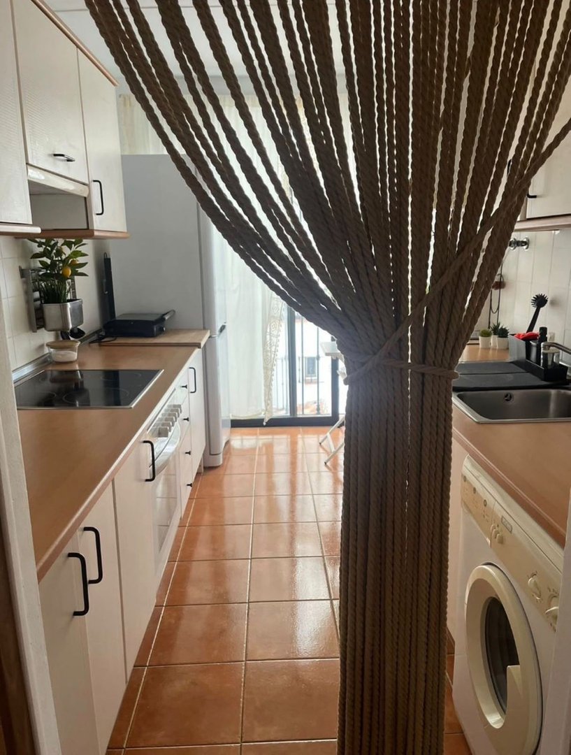 A kitchen with white cabinets, a stovetop, and a rope curtain in the foreground. There is a washing machine and a sink visible.