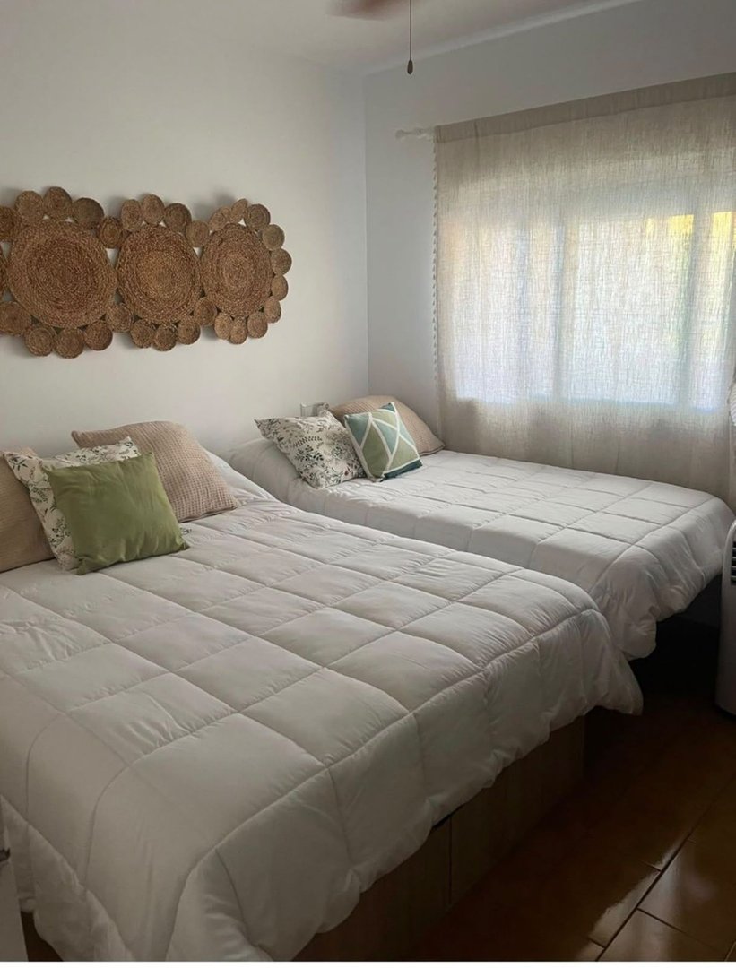 A bright bedroom with two beds, decorative jute wall art, and linen curtains filtering natural light.