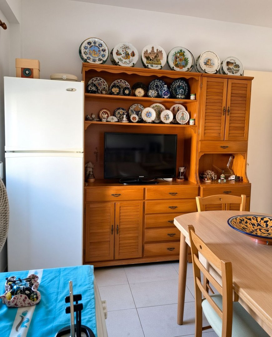 A kitchen with a wooden hutch displaying decorative plates, a television, a refrigerator, and a dining table with chairs.