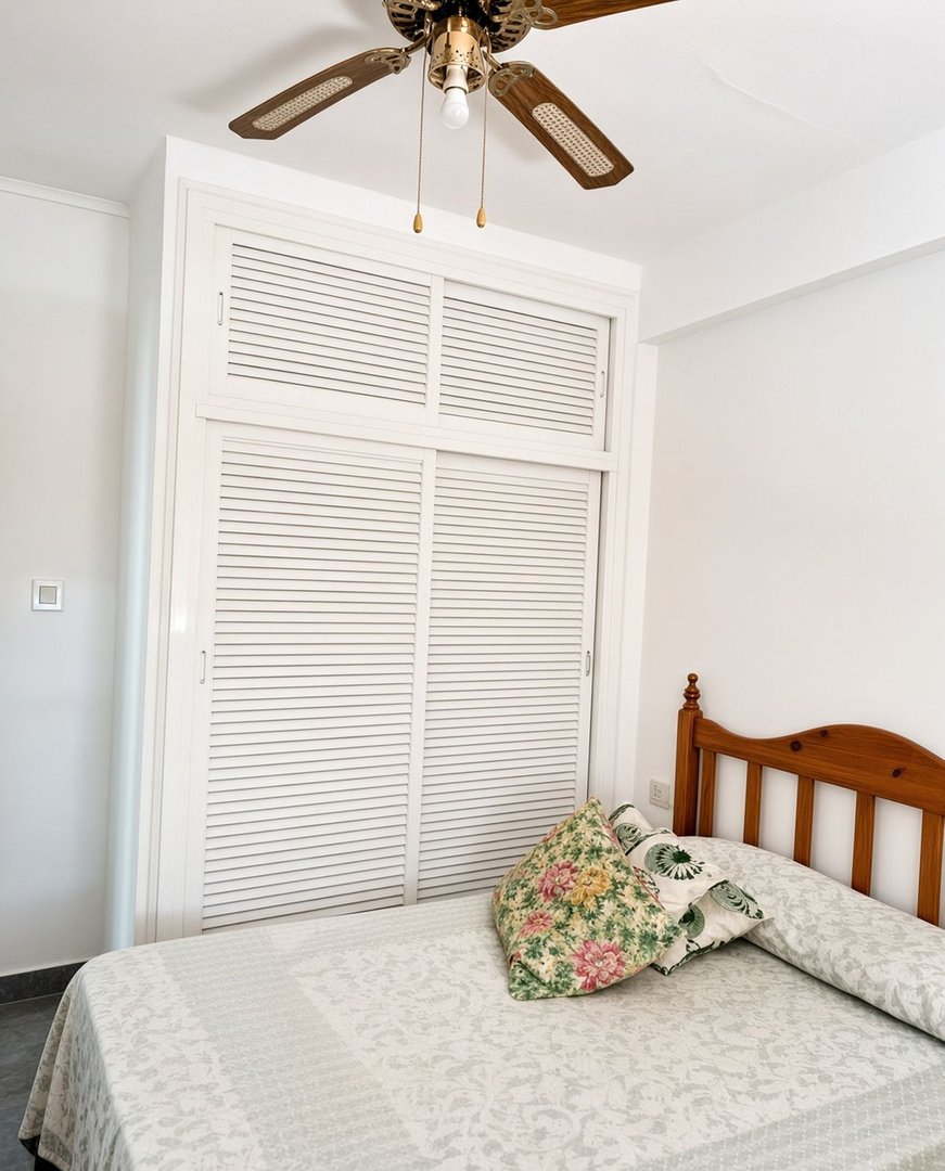 A bedroom with a bed, floral pillows, a white louvered wardrobe, and a ceiling fan overhead.