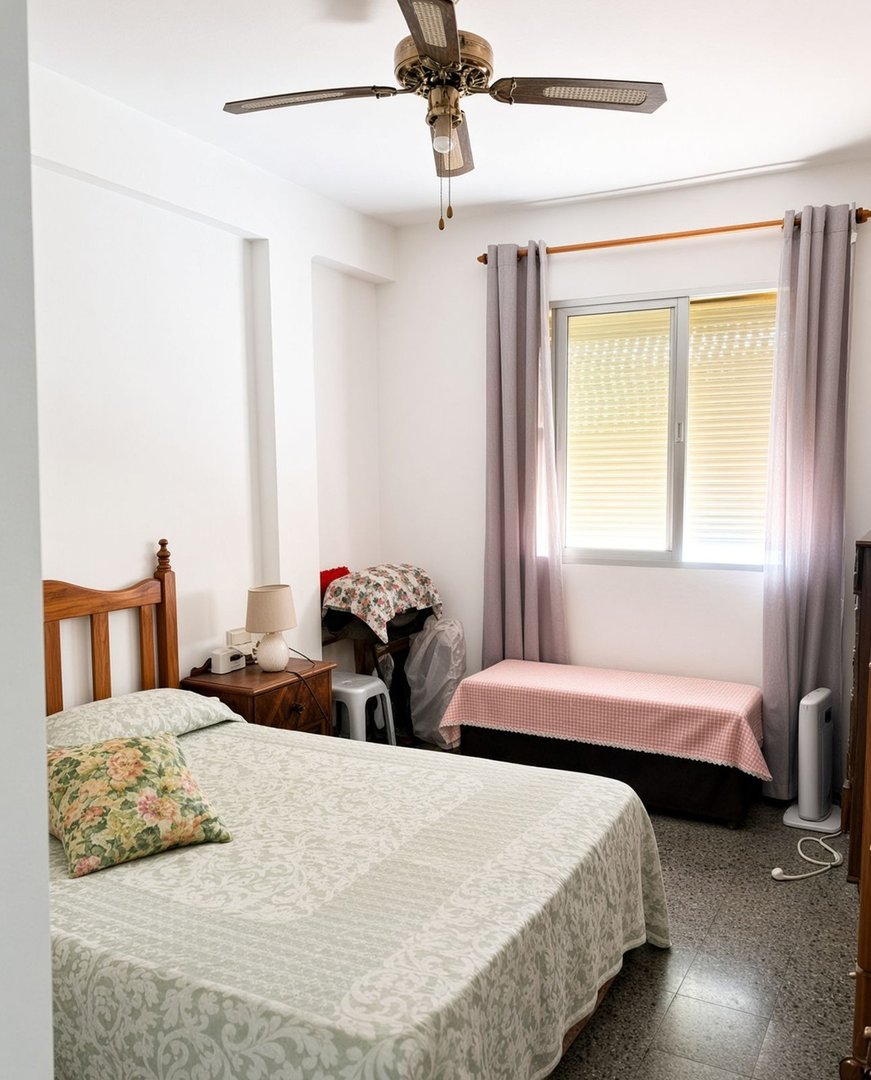 A bedroom with a bed, bedside table, ceiling fan, and a window with grey curtains.