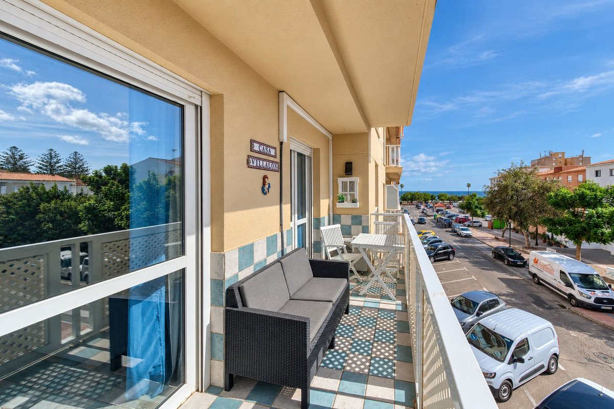 A sunny balcony with seating overlooking a street with cars and the sea in the distance.