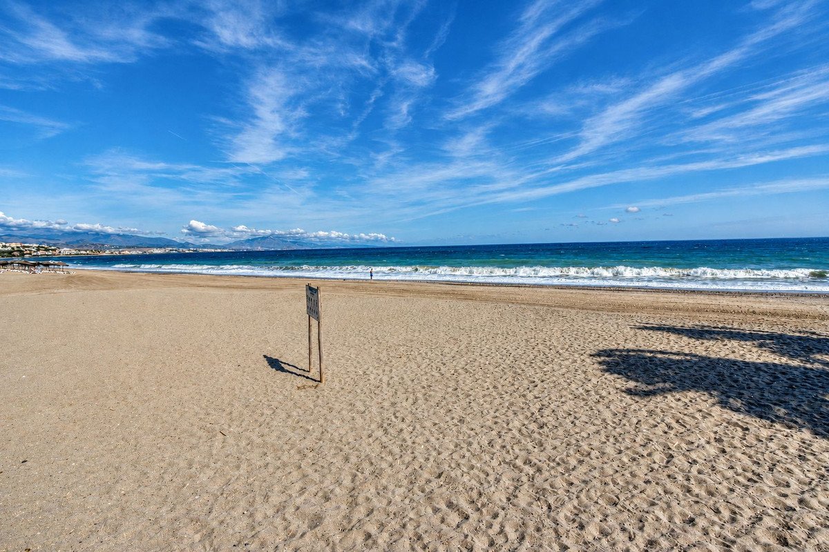 A wide sandy beach with gentle waves and a distant coastline under a blue sky with wispy clouds.