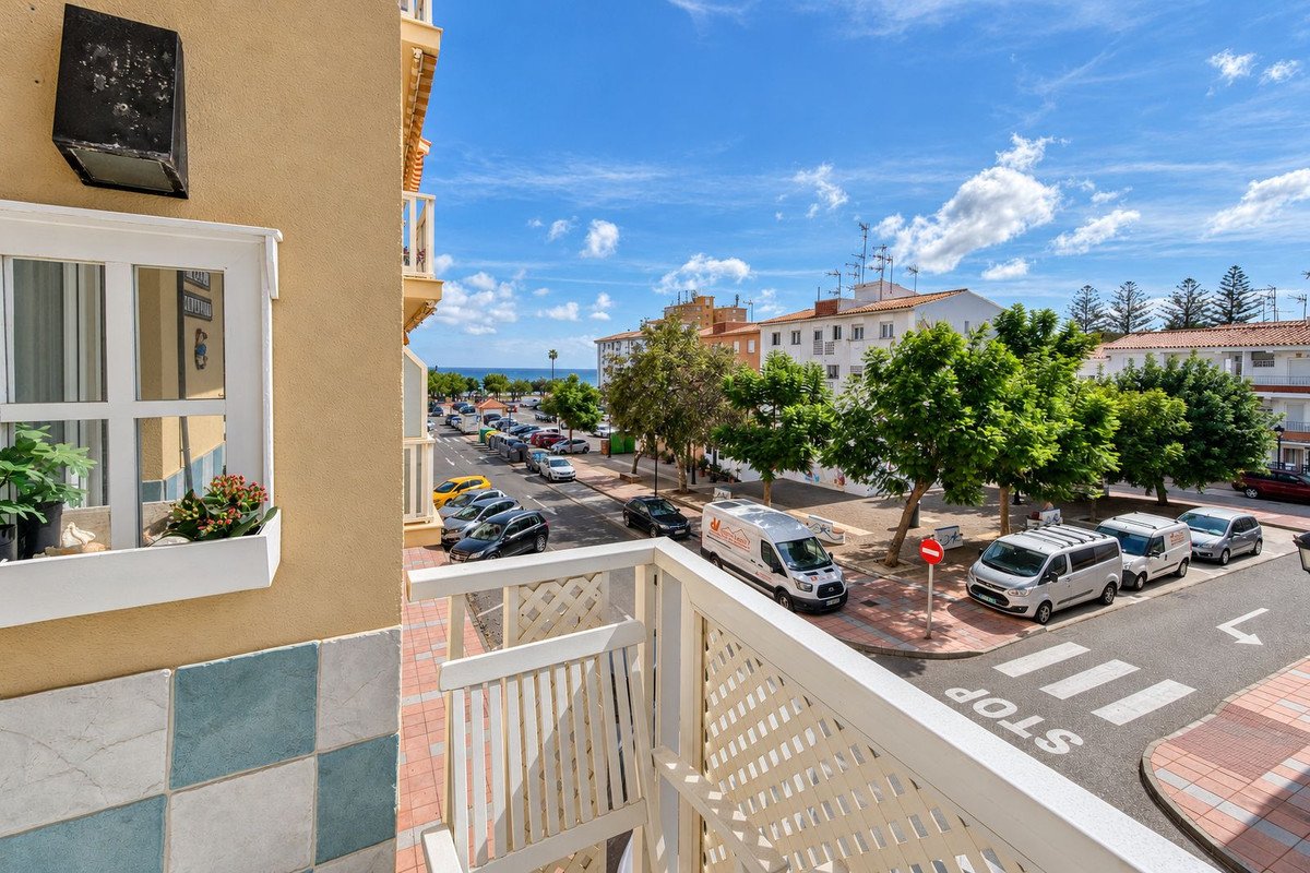 View from a balcony overlooking a street with cars, trees, buildings, and the sea in the distance under a blue sky.