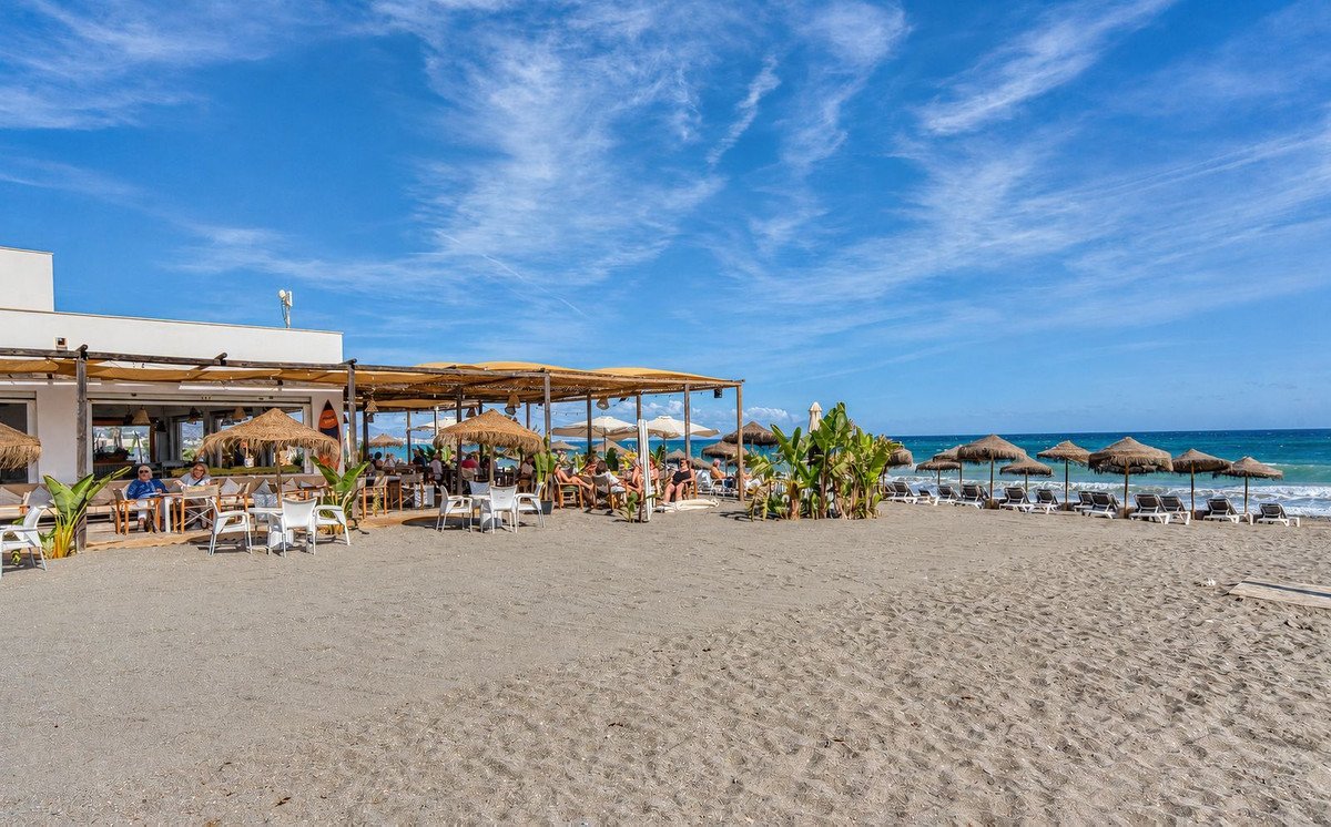 Beach restaurant with tables and umbrellas on the sand, overlooking the sea under a blue sky with clouds.
