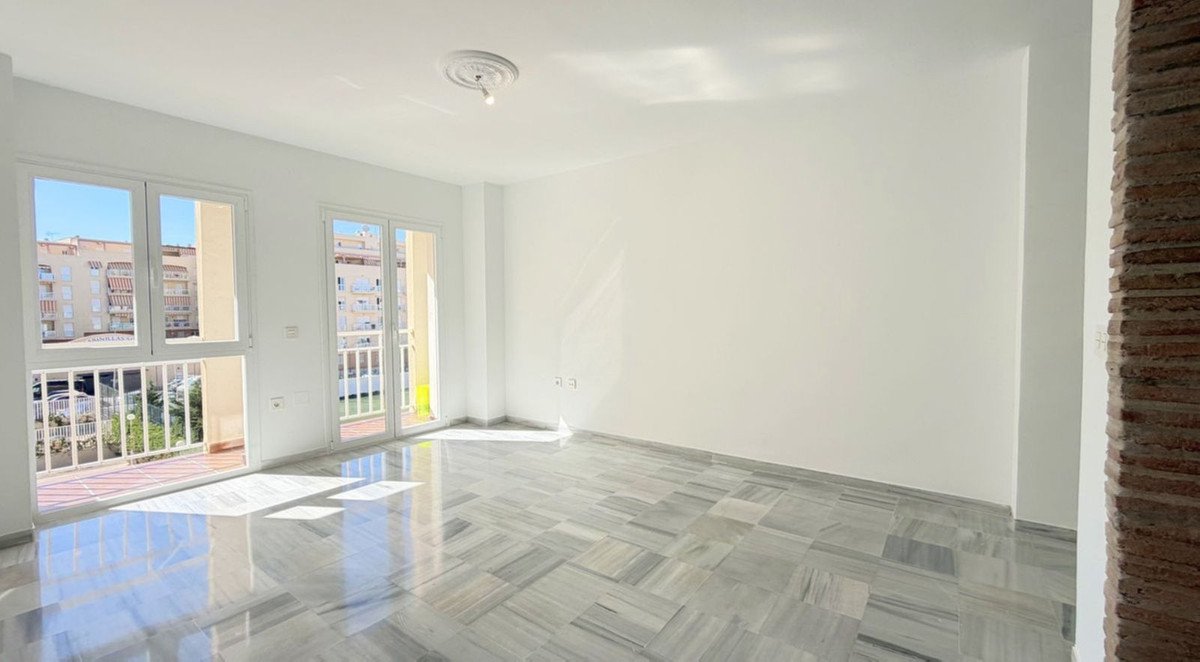 Empty living room with white walls, marble floors, and large windows leading to a balcony with an outdoor view.
